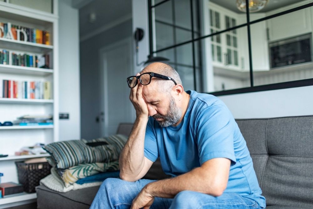 A middle-aged man sits on a couch, head in hand, looking stressed or upset. Glasses rest on his head. There are bookshelves and a modern interior in the background.