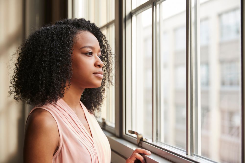 A woman with curly hair wearing a sleeveless pink top gazes thoughtfully out of a large window, with soft daylight illuminating her face.