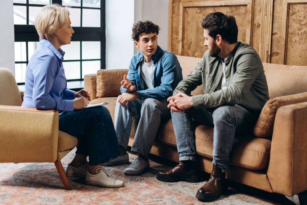 Three people sit in a living room: a woman with a notebook, a young man in a blue shirt speaking, and an older man listening. They appear engaged in a serious conversation.