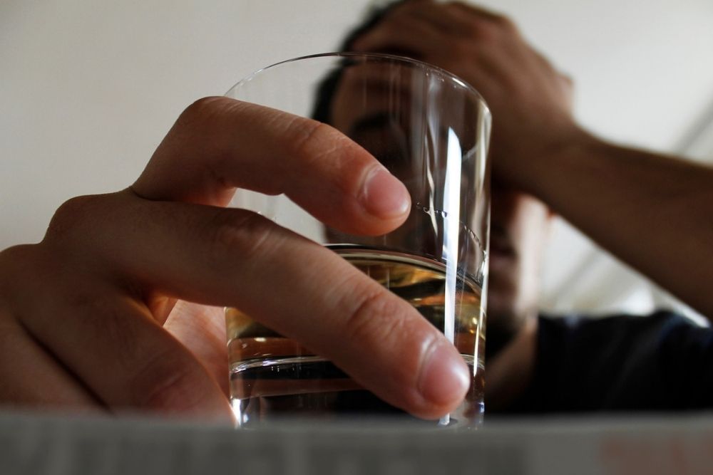 A person holds a glass of clear liquid, possibly water or alcohol, with one hand while the other hand covers part of their face, suggesting stress or contemplation. The background is out of focus.