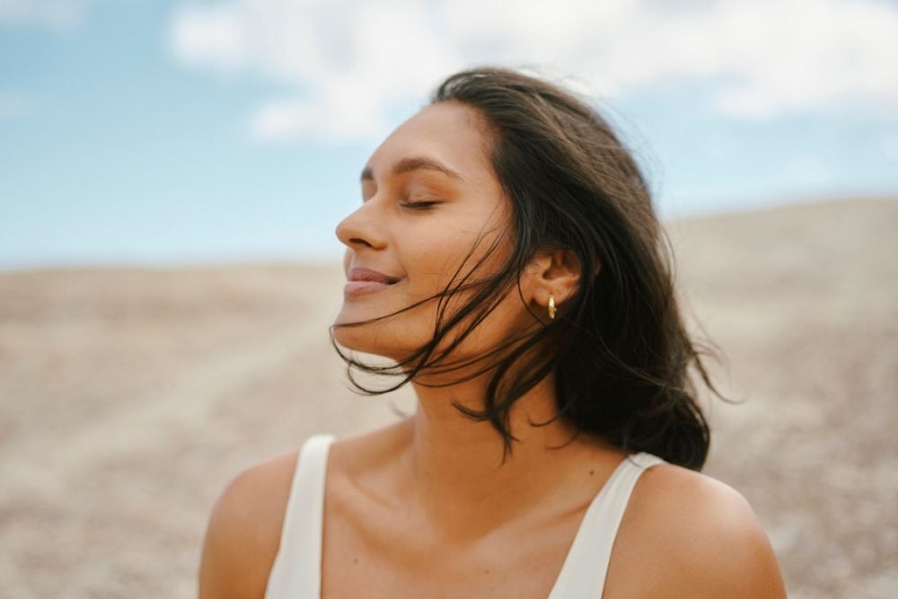 A woman with closed eyes and a gentle smile stands outdoors, wind blowing her dark hair. She wears a white top and gold hoop earrings, with a blurred natural landscape and blue sky with clouds in the background.