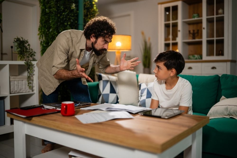 A man gestures intensely while talking to a boy sitting at a table with papers and a tablet. The boy looks up at him, appearing upset. They are in a cozy living room with a green couch and shelves in the background.