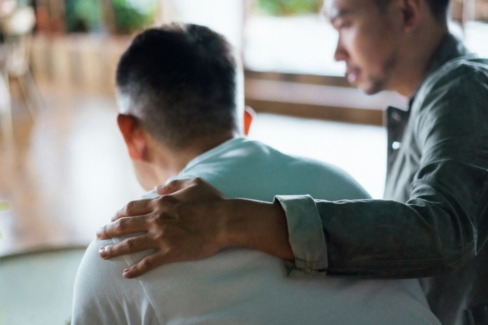 Two people sit together indoors, one with a hand on the others shoulder, offering comfort or support in a blurred, softly lit setting.