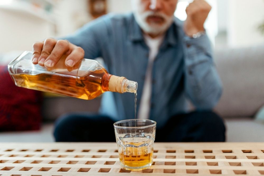 A man with a gray beard pours whiskey from a bottle into a glass while sitting at a wooden table.