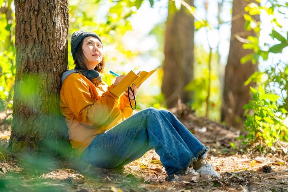 A person in a yellow jacket and beanie sits against a tree in a forest, writing in a notebook. Headphones rest around their neck, and sunlight filters through the leaves.