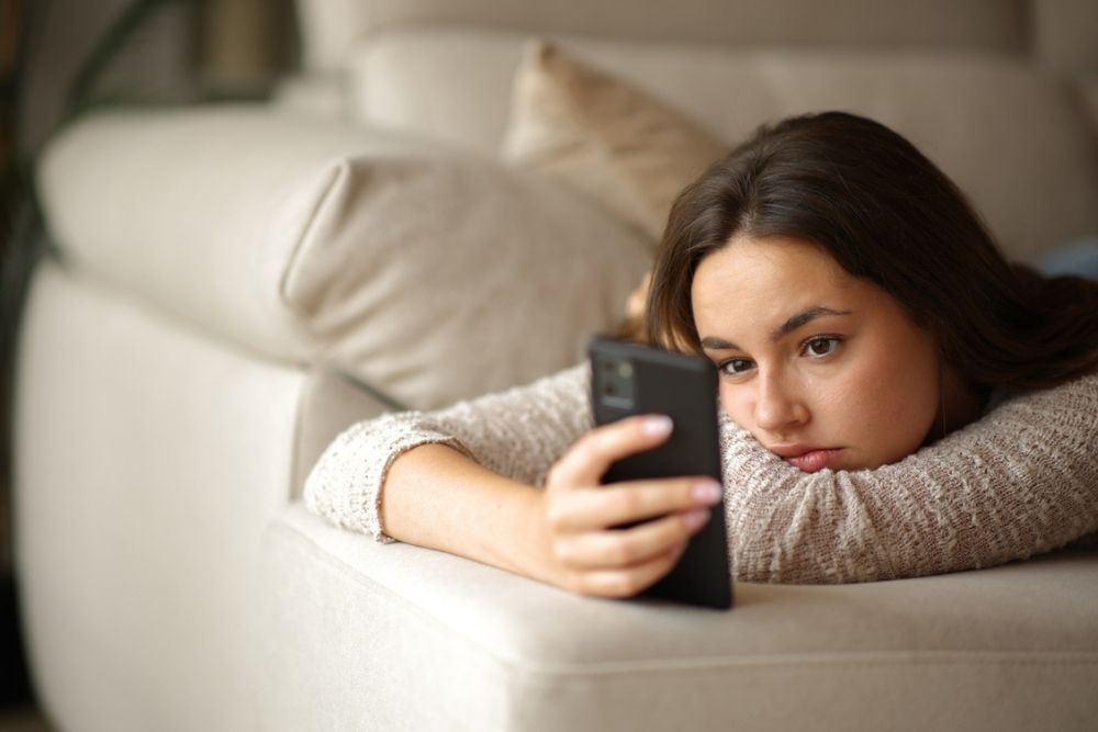 A young woman lies on a couch, resting her head on her arm and looking thoughtfully at a smartphone she is holding in her hand.