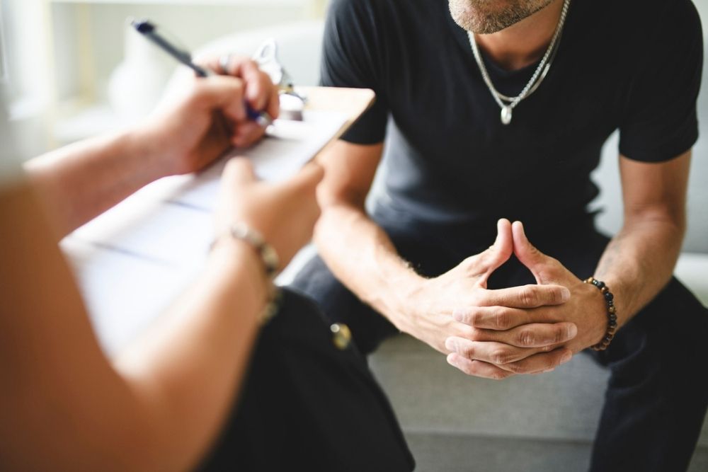 A man sits with his hands clasped while another person writes on a clipboard, suggesting a counseling or therapy session.