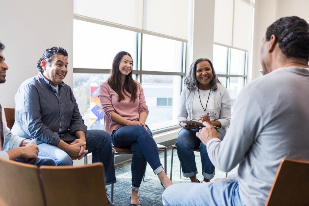 Five people sit in a circle talking and smiling in a bright room with large windows; one person holds a notepad, suggesting a group discussion or support meeting.