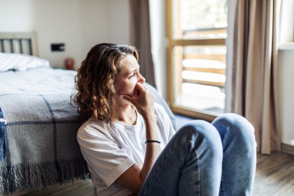 A woman sits on the floor by her bed, looking thoughtfully out a window, resting her hand on her mouth.