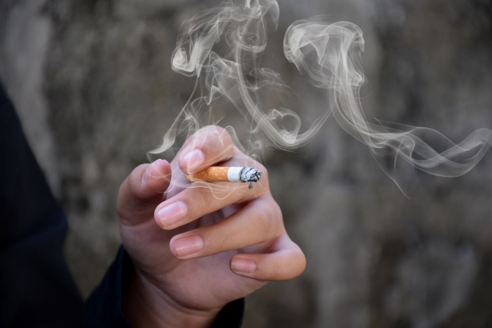 A hand holding a lit cigarette with swirling smoke against a blurred gray background.