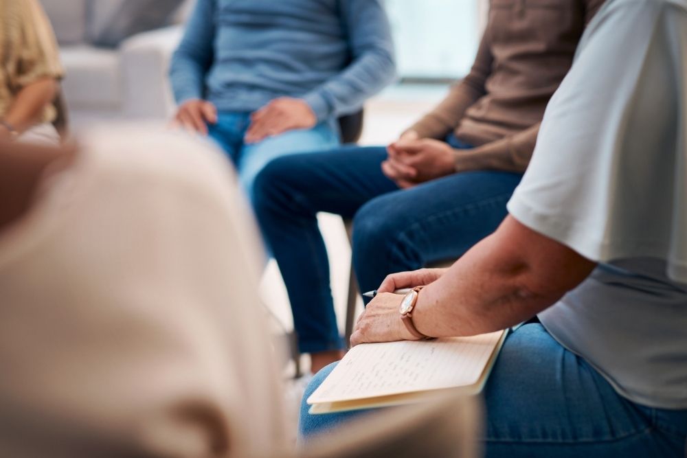 Several people sit in a circle, wearing casual clothes. One person in the foreground holds a notepad and pen, suggesting a group discussion or support meeting setting. Faces are not visible.