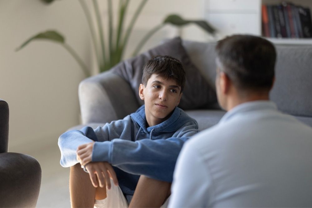 A teenage boy sits on the floor, looking thoughtfully at an adult man sitting nearby in a living room with a sofa and plants in the background.