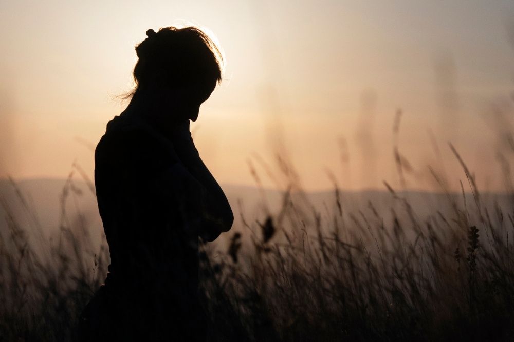 Silhouette of a person standing in tall grass at sunset, with head slightly bowed and hands near the chin, creating a calm and contemplative mood.