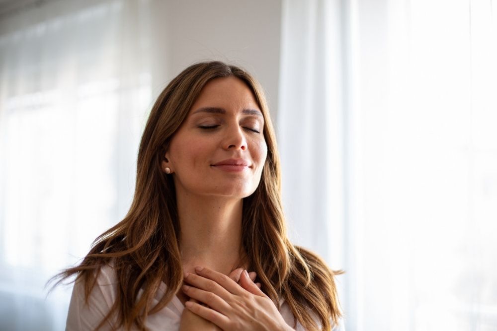 A woman with long brown hair stands indoors with her eyes closed and both hands over her chest, appearing calm and content. Soft daylight comes through sheer curtains in the background.