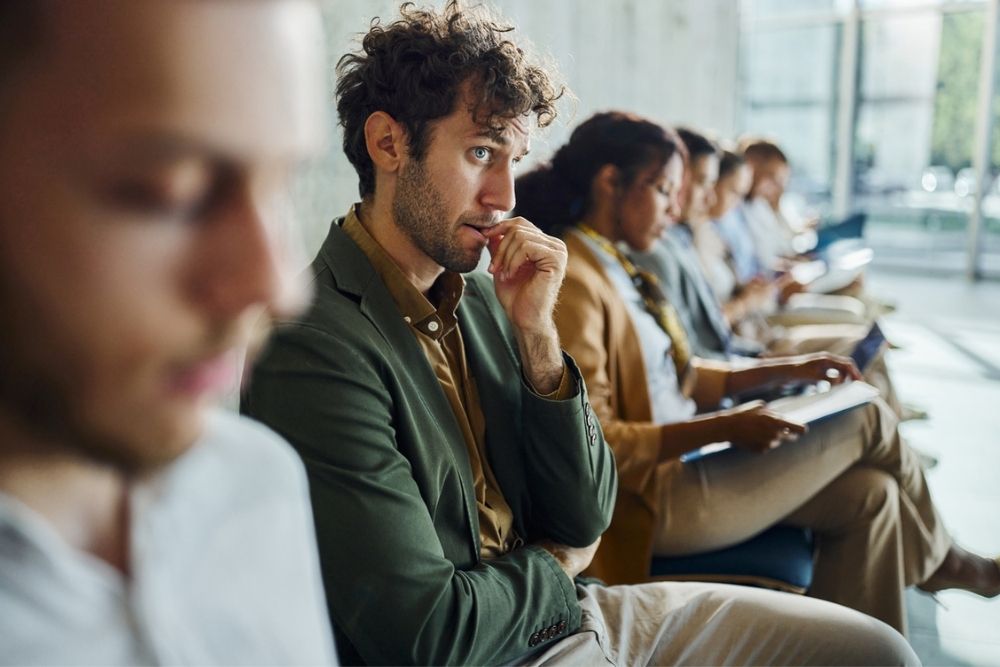 A row of people sitting and waiting. One man in focus, wearing a green jacket, looks anxious and bites his fingernail. Others appear to be waiting patiently, looking forward or down, some with papers in hand.