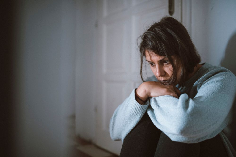 A woman sits on the floor indoors with her arms around her knees, looking down with a sad and thoughtful expression.