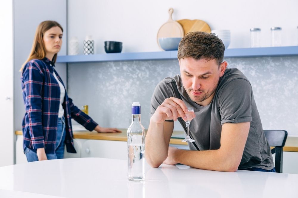 A man sits at a kitchen counter holding a glass of clear liquid, looking down. An unhappy woman stands in the background, watching him. A bottle sits on the counter between them.