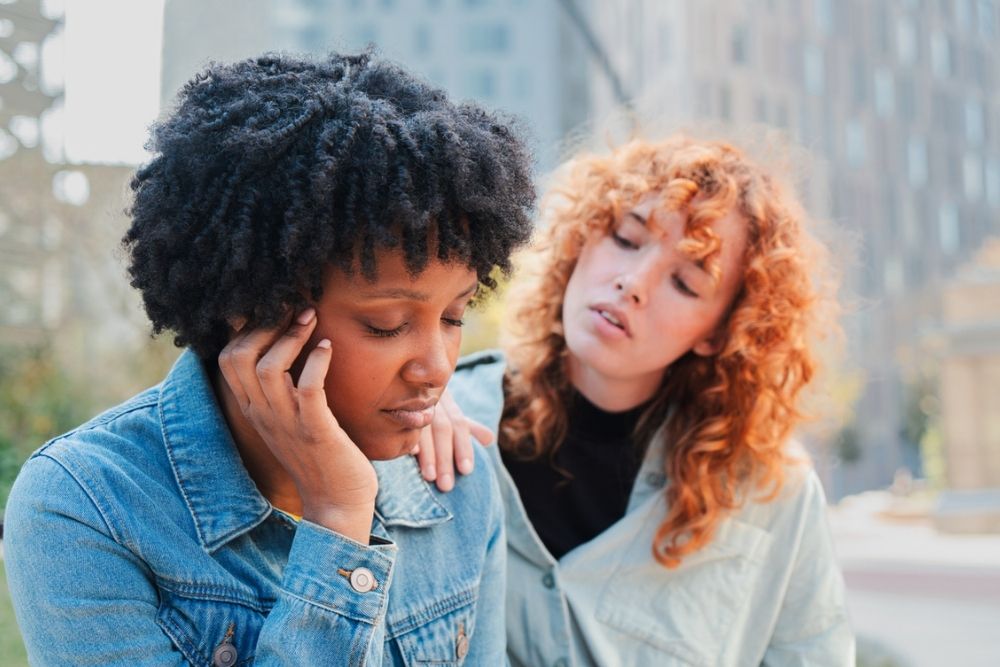 Two women sit outdoors; one appears sad with her hand on her face, while the other gently touches her shoulder, offering comfort. Both wear denim jackets and have curly hair, with blurred buildings in the background.