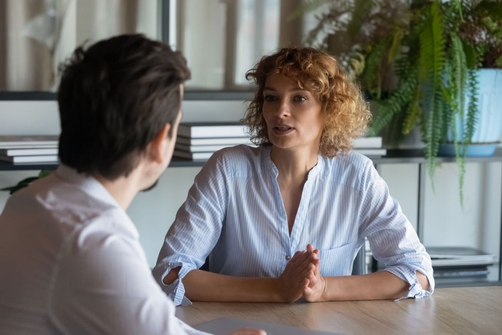 Two people sit across from each other at a table, engaged in conversation. The woman speaks with her hands clasped, while the man listens attentively. There are plants and shelves with books in the background.