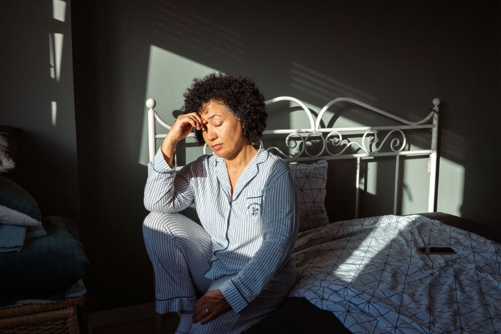 A woman in striped pajamas sits on the edge of her bed with her eyes closed, resting her hand on her forehead. Sunlight streams through a window, casting shadows on the wall and bed.