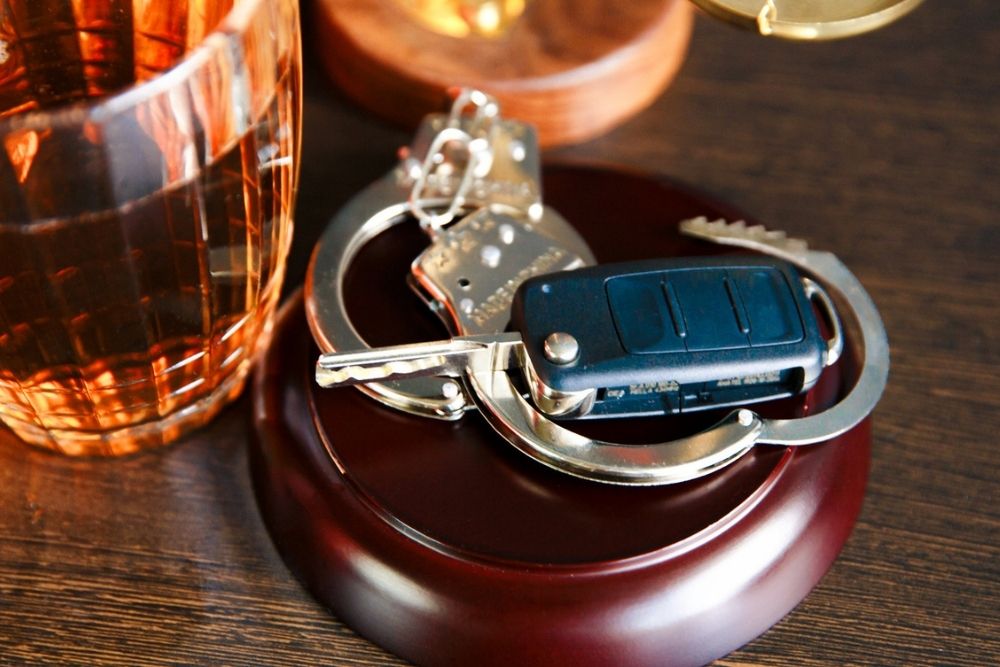 A set of car keys and handcuffs resting on a wooden surface next to a glass of amber liquid, possibly whiskey, in a faceted glass.