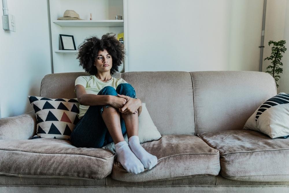 A woman with curly hair sits on a beige couch, hugging her knees, wearing a light shirt, jeans, and socks. Two patterned pillows are beside her, and shelves are visible in the background.