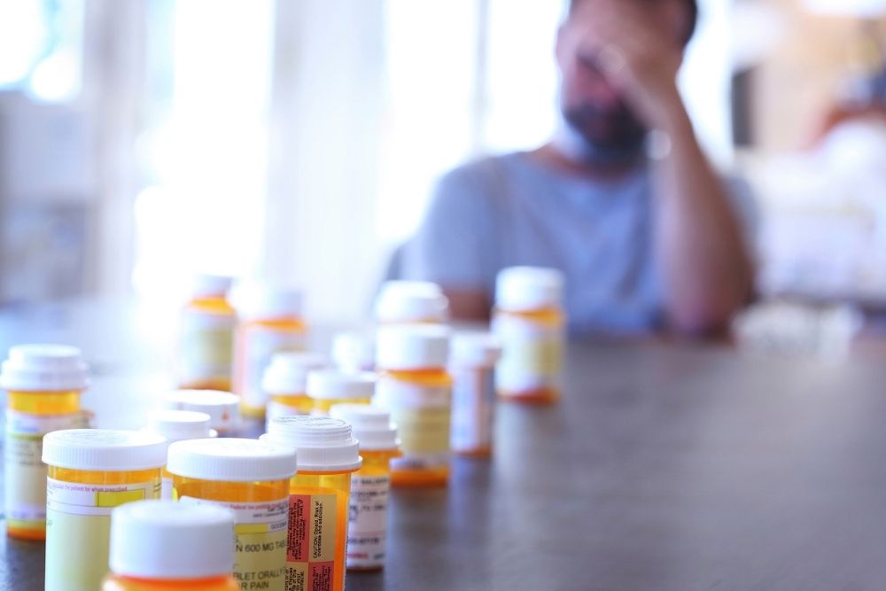A blurred person sits at a table with numerous prescription pill bottles in focus in the foreground, suggesting themes of medication, health, or mental health struggles.