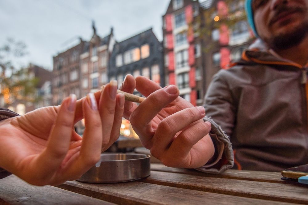 Two people sit at a wooden table outdoors, passing a rolled cigarette or joint between them. An ashtray is on the table, and colorful buildings are blurred in the background.