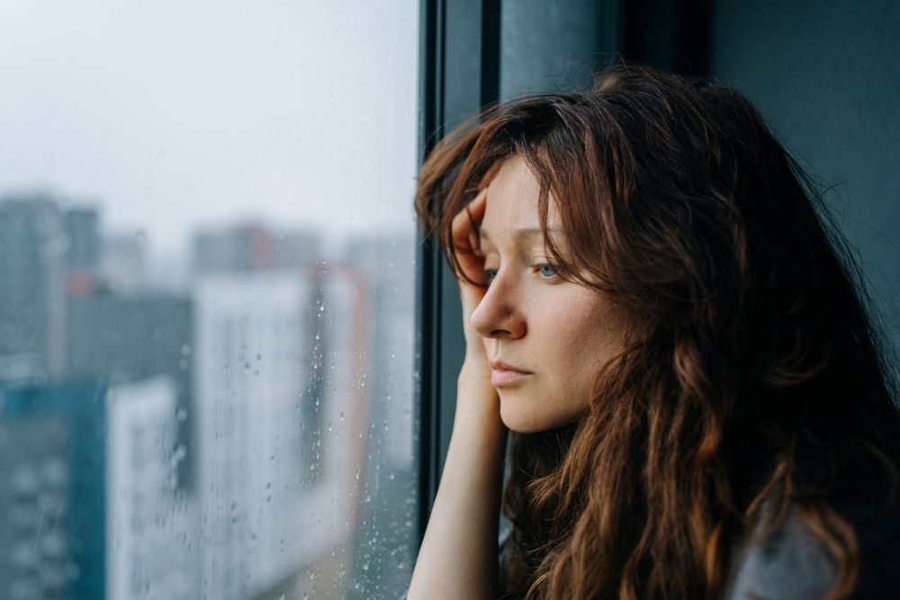 A woman with long brown hair looks out a window on a rainy day, resting her head on her hand, appearing thoughtful or sad. City buildings are blurred in the background.