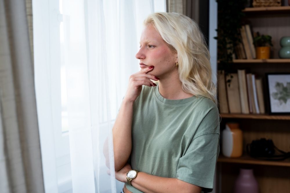 A woman with long blonde hair stands by a window, looking outside thoughtfully with her hand on her chin. She wears a green shirt and a watch, with books and plants on shelves in the background.
