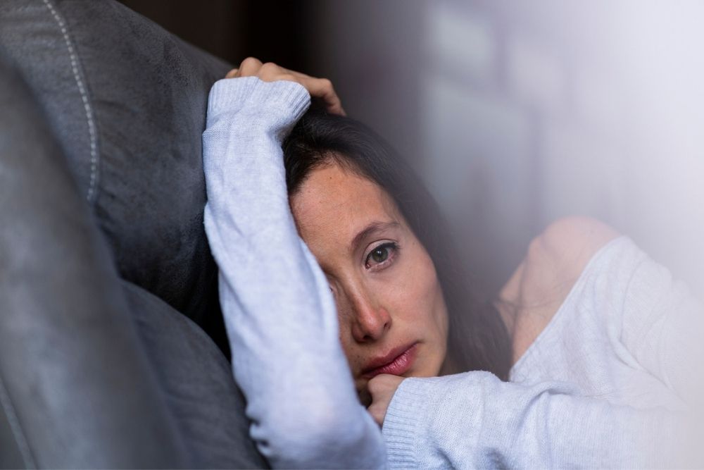 A woman with long dark hair lies on a couch, resting her head on her arm and looking sad or thoughtful. She wears a light sweater and gazes directly at the camera.