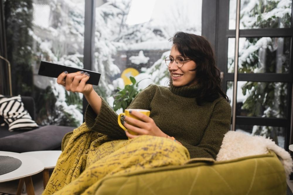 A person in glasses sits on a chair with a yellow blanket, holding a yellow mug and a remote, smiling. Snowy trees are visible through large windows in the background.