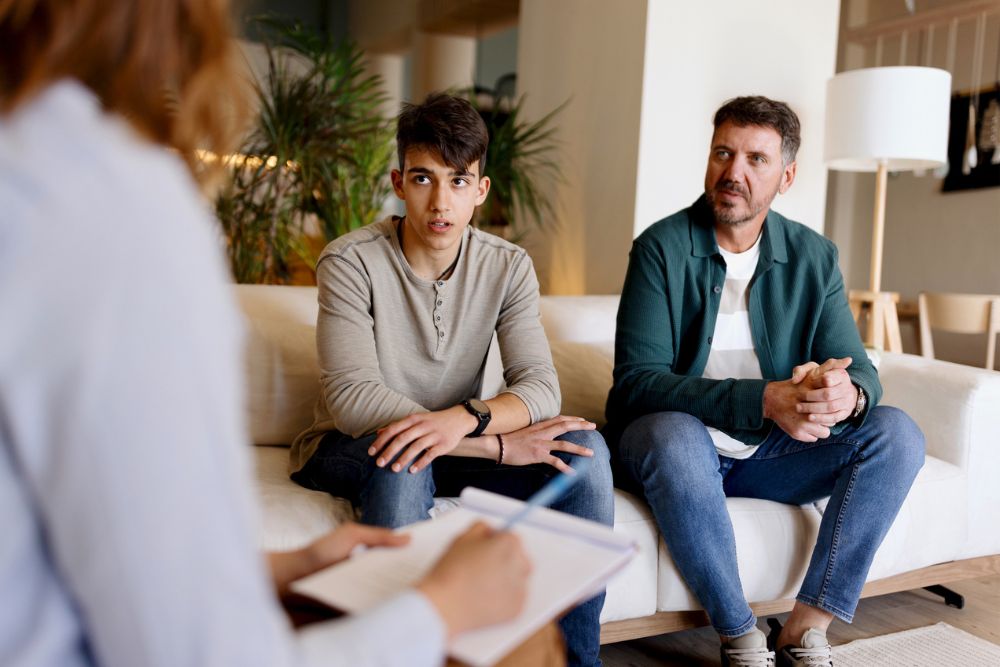 A young man and an older man sit on a couch, facing a person taking notes. The setting appears to be a therapy or counseling session in a comfortable, modern room.