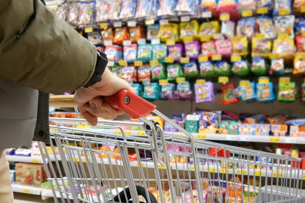 A person pushes a shopping cart down an aisle filled with colorful packaged goods in a grocery store.