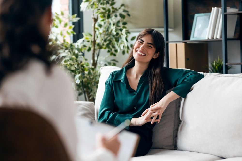 A smiling woman sits on a couch, talking to another person whose back is to the camera, in a bright room with plants and shelves in the background.