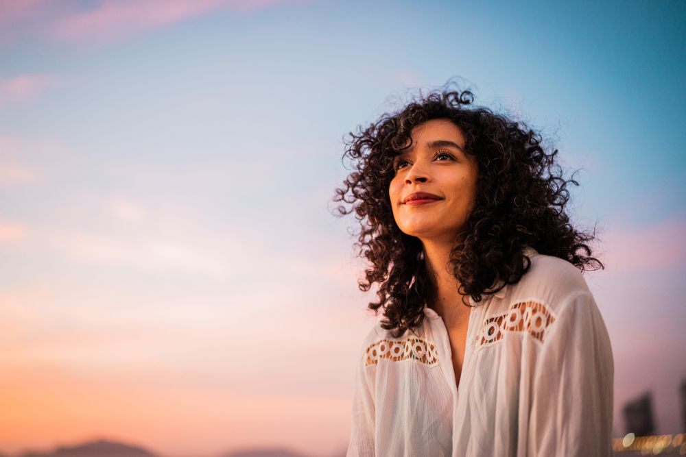 A woman with curly hair and a white blouse smiles gently while looking up, standing outdoors at sunset with a colorful sky in the background.