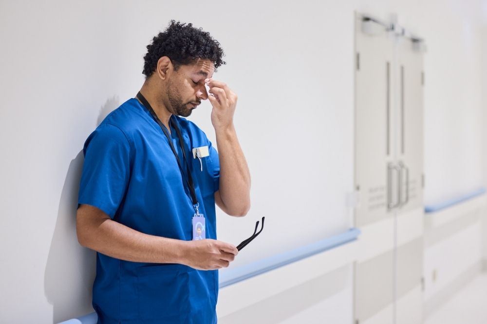A healthcare worker in blue scrubs stands in a hallway, holding glasses and rubbing his eyes, appearing tired or stressed.