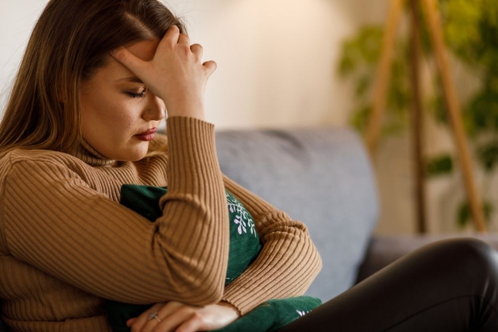 A woman sits on a couch with her eyes closed, holding her forehead and hugging a pillow, appearing stressed or upset.