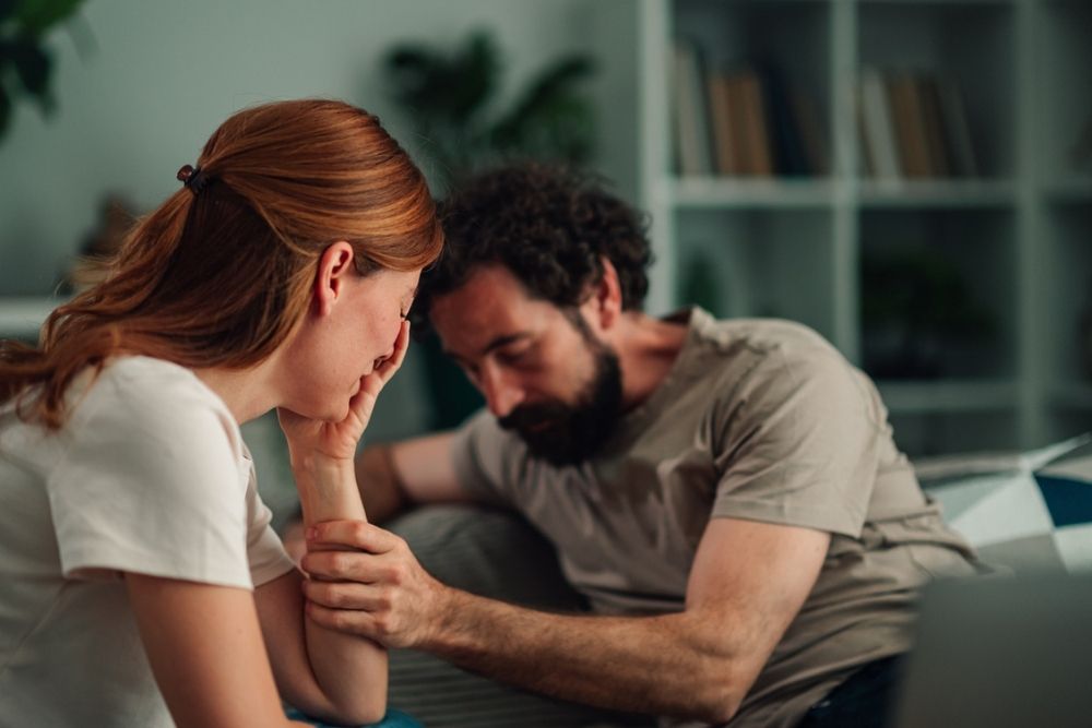 A man comforts a woman who appears upset, holding her arm as she sits with her head in her hand. They are indoors, sitting closely together on a couch.