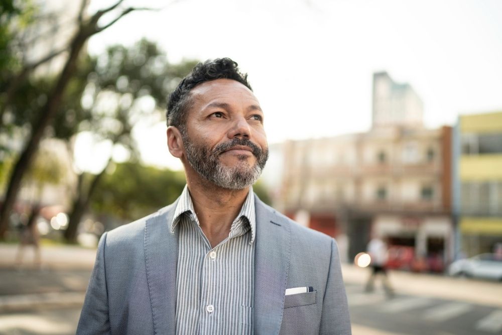A man with short curly hair and a beard, wearing a suit, stands outdoors on a sunny day, looking off into the distance with a slight smile. Blurred city buildings and trees are visible in the background.