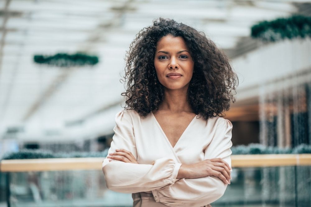 Woman with curly hair stands indoors with arms crossed, wearing a light-colored blouse, looking confidently at the camera. The background is modern and softly blurred.