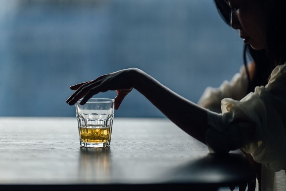 A person sitting at a table, reaching for a glass filled with a golden liquid, with a moody and dimly lit background.