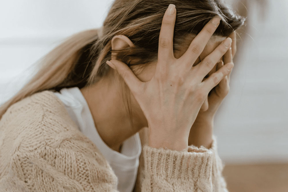 A person with light brown hair and a beige sweater sits with their head in their hands, appearing stressed or upset.