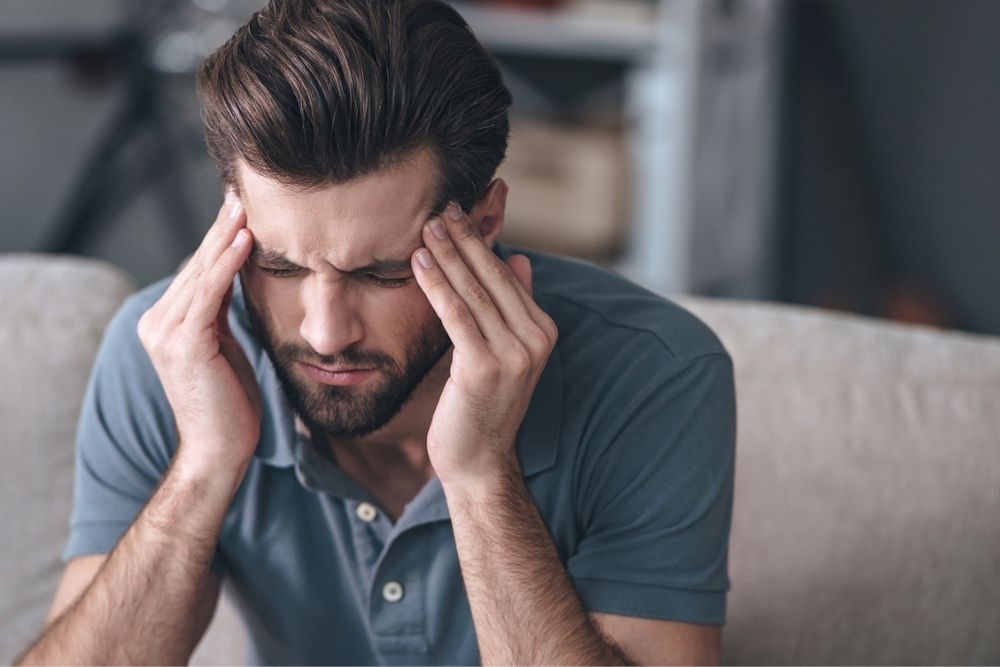 A man with brown hair and a beard sits on a couch with his eyes closed, pressing his fingers to his temples, appearing to have a headache or to be stressed.