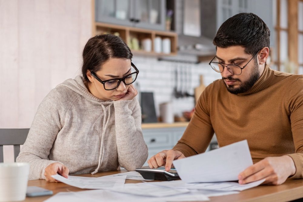 A worried couple sits at a table reviewing bills and paperwork, with a calculator and documents spread out in front of them in a kitchen setting.
