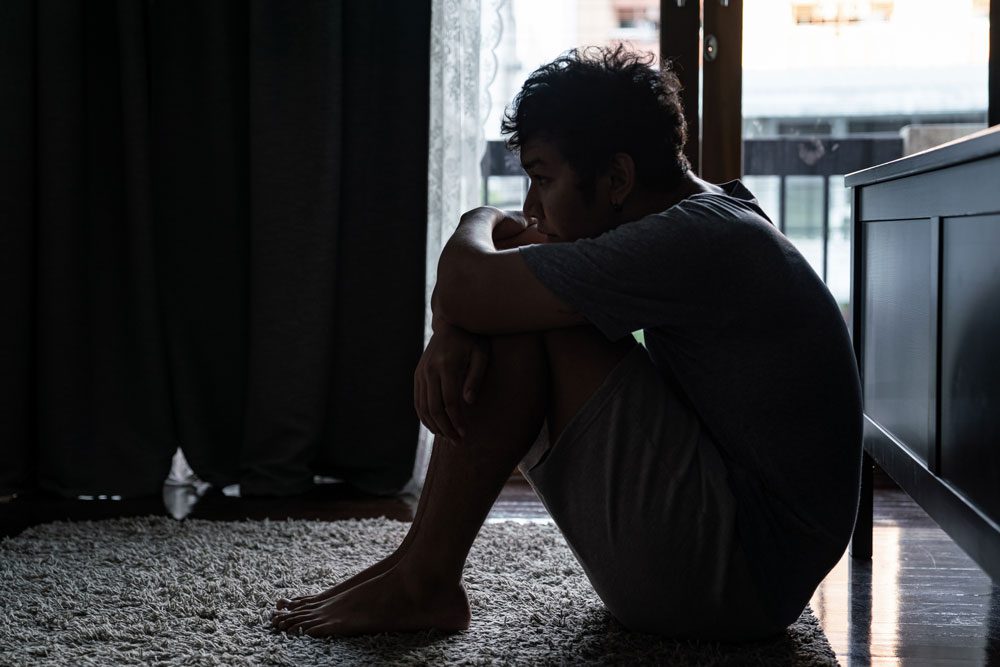 A person sits on the floor in a dimly lit room, hugging their knees and looking out toward a window, appearing deep in thought or possibly sad.