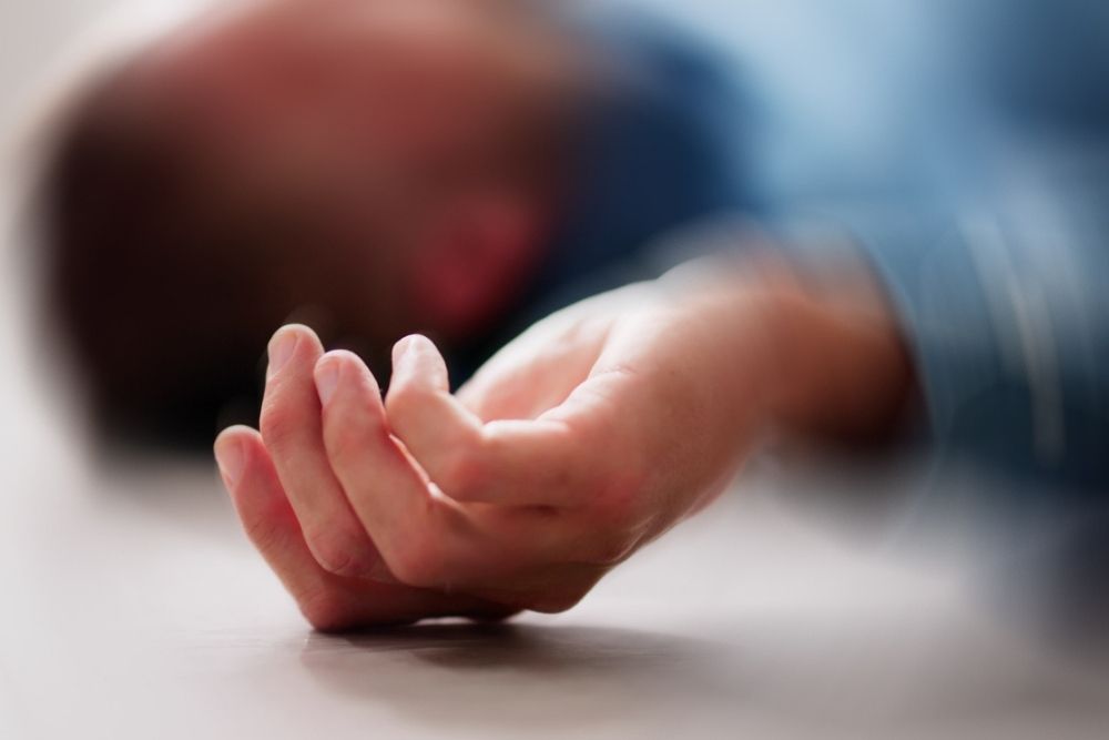 A person lies on the ground with a hand in focus and the face blurred, suggesting unconsciousness or distress. The individual wears a blue shirt.