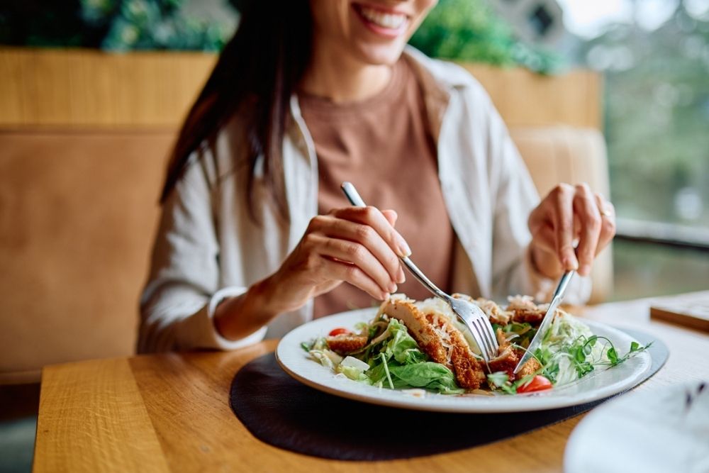 A woman smiles while cutting into a salad with crispy chicken at a restaurant, sitting at a wooden table with greenery visible in the background.