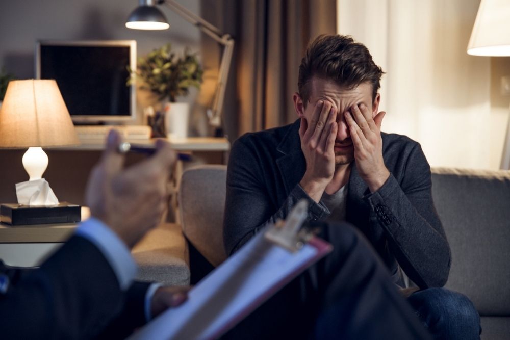 A distressed man sits on a couch covering his face with his hands while another person, possibly a therapist, holds a clipboard and pen during a conversation in a warmly lit room.