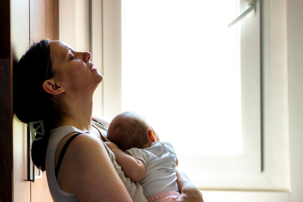 A tired woman with closed eyes leans against a wall, holding a baby in her arms, bathed in natural light from a nearby window.